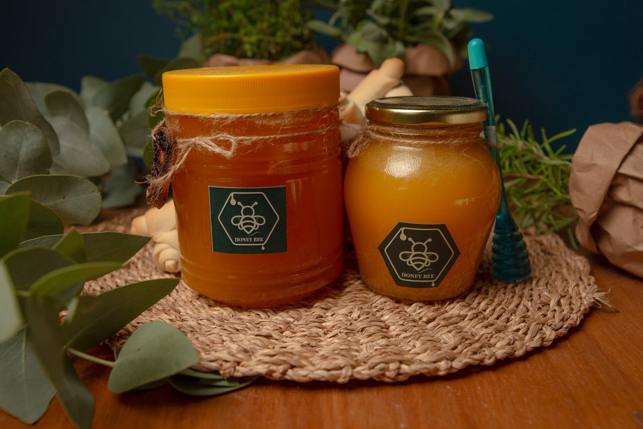 Two jars of honey on a woven mat surrounded by eucalyptus leaves and herbs.